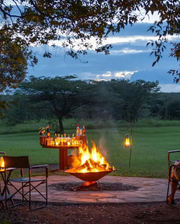 Guests of Encounter Mara Camp seated around an open campfire at dusk, enjoying drinks and lantern light overlooking open savannah and trees.