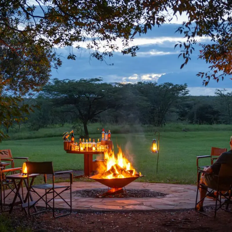Guests of Encounter Mara Camp seated around an open campfire at dusk, enjoying drinks and lantern light overlooking open savannah and trees.