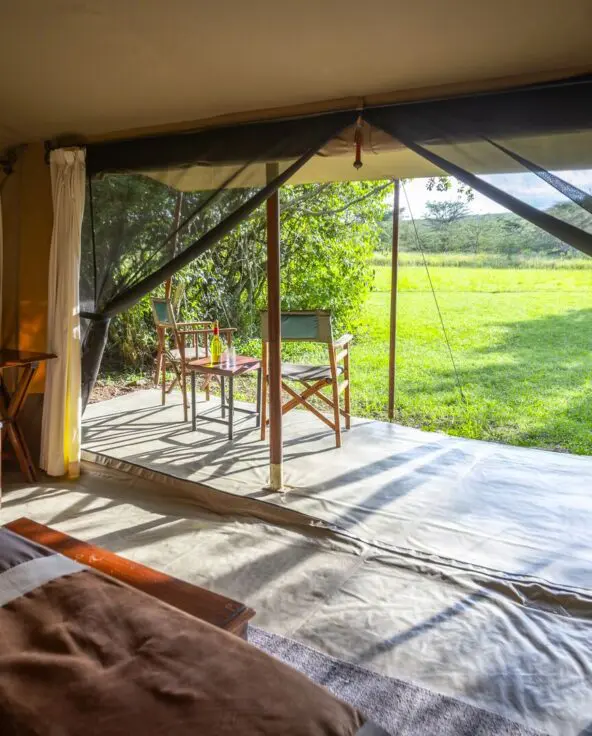 Interior of a safari tent with bed and open front leading to a grassy outdoor seating area