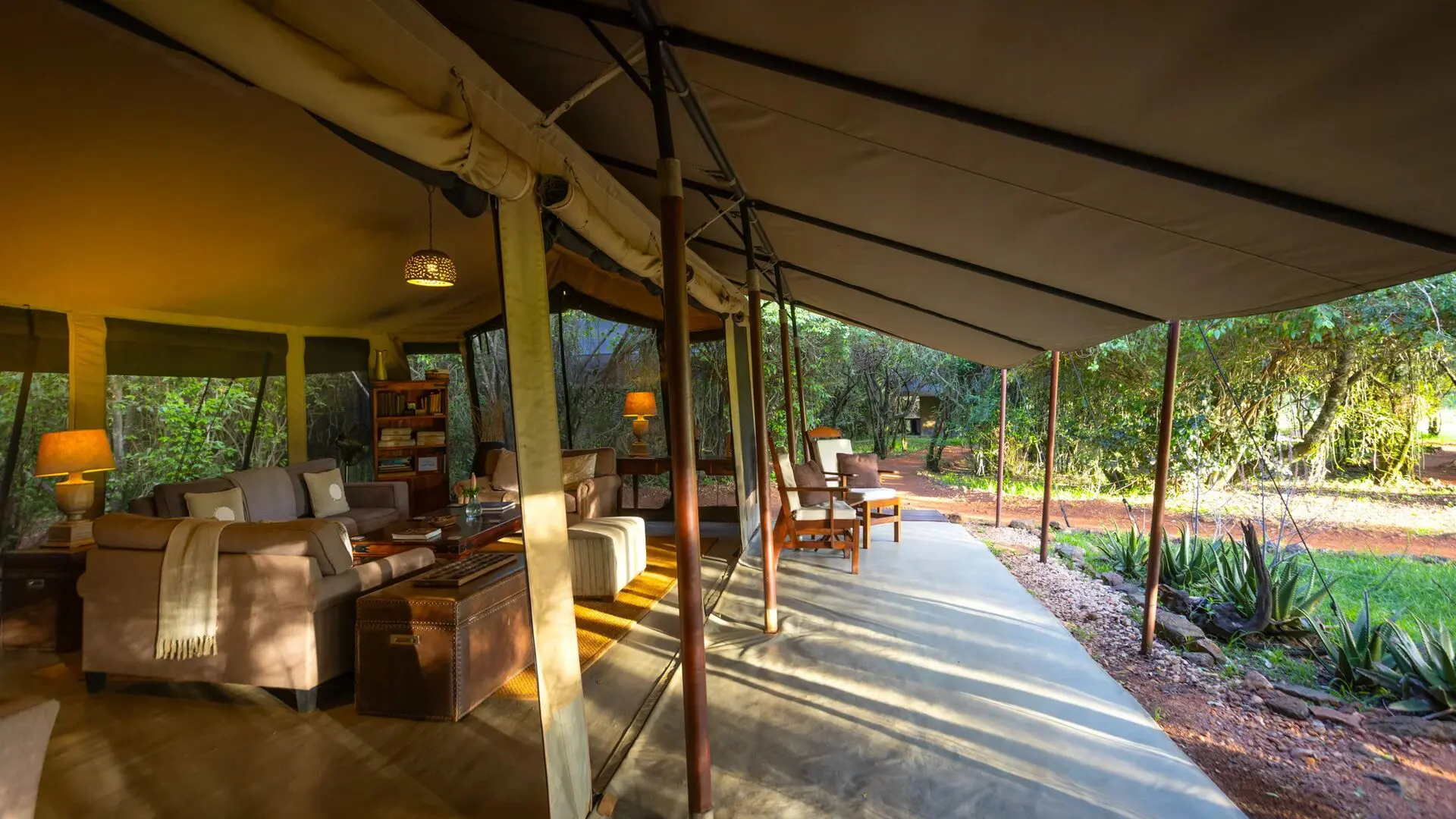 Interior of Encounter Mara's luxury safari tent lounge with sofas, lamps, wooden furniture, and an open veranda overlooking forested surroundings.