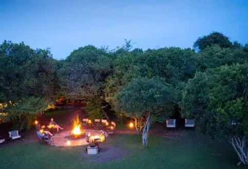 Aerial view of a safari camp at night with a central fire pit, lantern-lit seating, and surrounding trees under a deep blue sky.