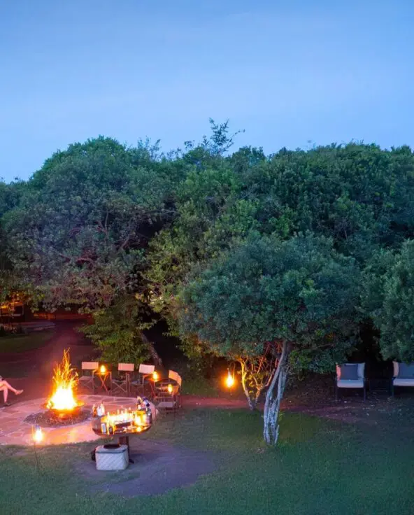 Aerial view of a safari camp at night with a central fire pit, lantern-lit seating, and surrounding trees under a deep blue sky.