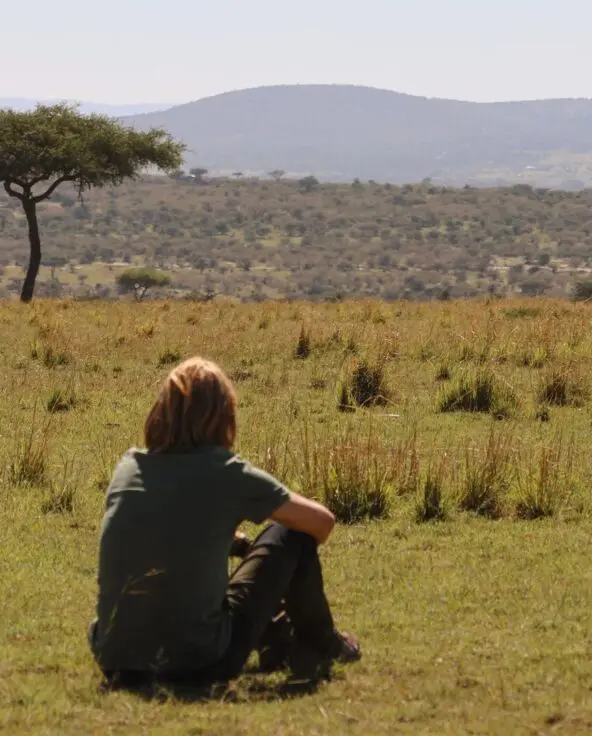 A person sitting on open grassland looking toward distant hills and a lone tree in the Maasai Mara