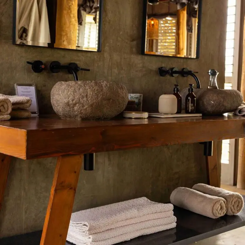 Jabali Private House bathroom with a wooden vanity, stone basin sinks, neatly folded towels, and natural light filtering through shuttered windows.