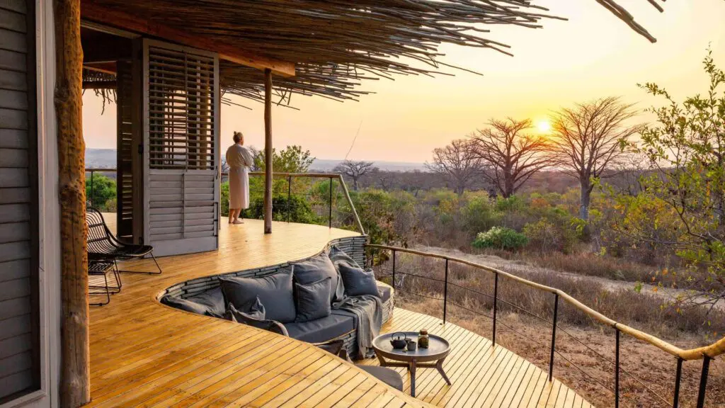 A guest stands on a private wooden deck at sunset, overlooking open woodland and baobab trees, with built-in seating and soft furnishings facing the view.