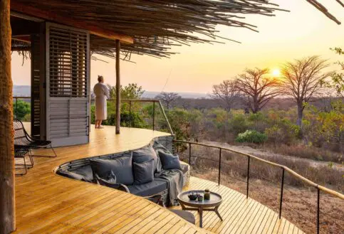 A guest stands on a private wooden deck at sunset, overlooking open woodland and baobab trees, with built-in seating and soft furnishings facing the view.
