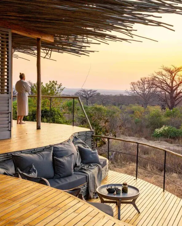 A guest stands on a private wooden deck at sunset, overlooking open woodland and baobab trees, with built-in seating and soft furnishings facing the view.