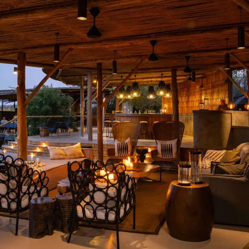 Jabali's open-air safari lodge lounge and bar at dusk, with wooden beams, warm lantern lighting, comfortable seating, and views over the surrounding bush.
