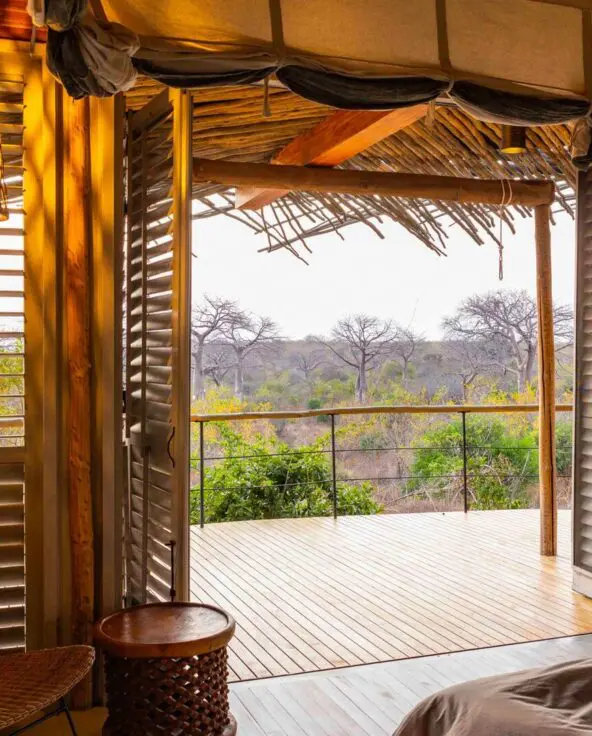interior of a suite at jabali camp with open wooden shutters leading onto a private deck, overlooking trees and open woodland, with soft natural light and woven furnishings inside