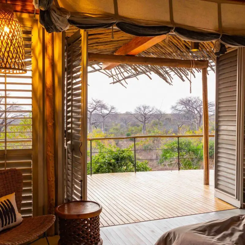 interior of a suite at jabali camp with open wooden shutters leading onto a private deck, overlooking trees and open woodland, with soft natural light and woven furnishings inside