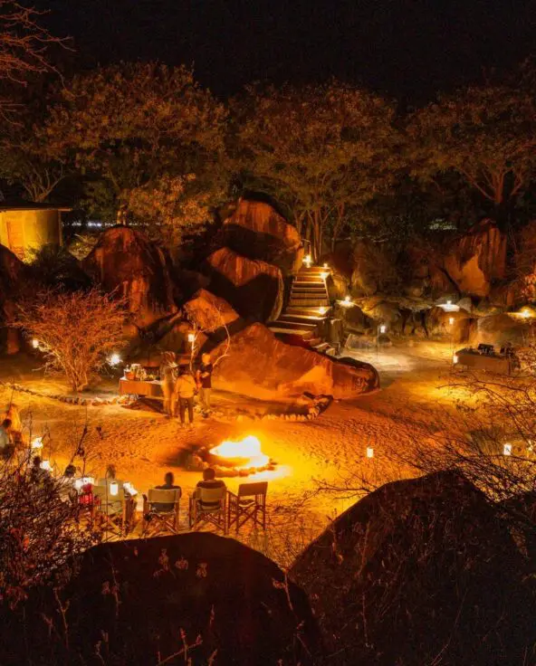 Guests gather around a fire pit at night, surrounded by softly lit boulders and trees, with lanterns illuminating an open-air lodge setting beneath the stars