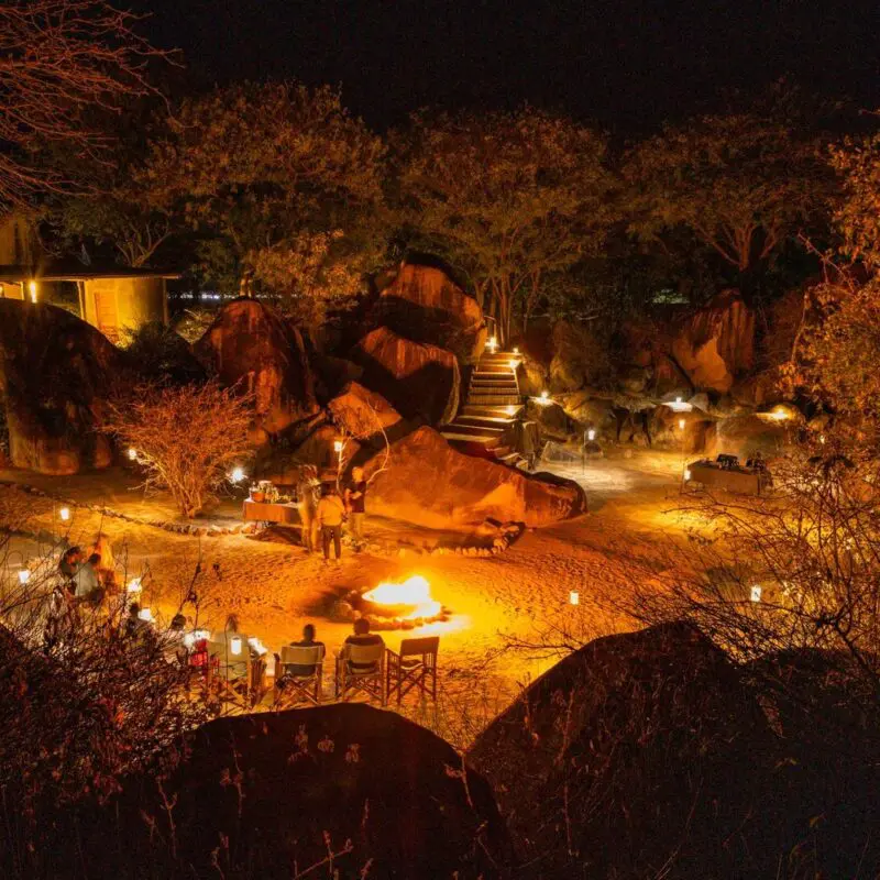 Guests gather around a fire pit at night, surrounded by softly lit boulders and trees, with lanterns illuminating an open-air lodge setting beneath the stars