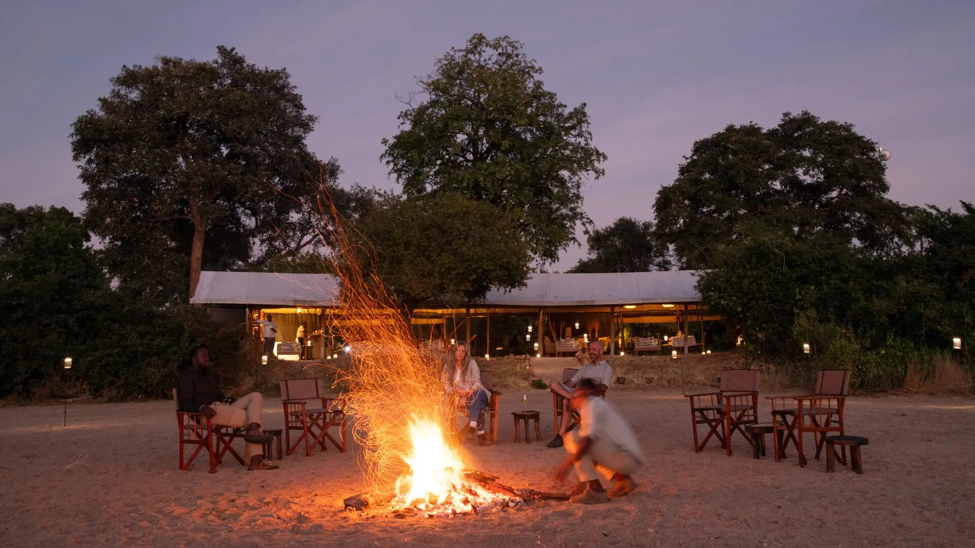 Kokoko Camp guests sit around the campfire at dusk with glowing tents and trees in the background