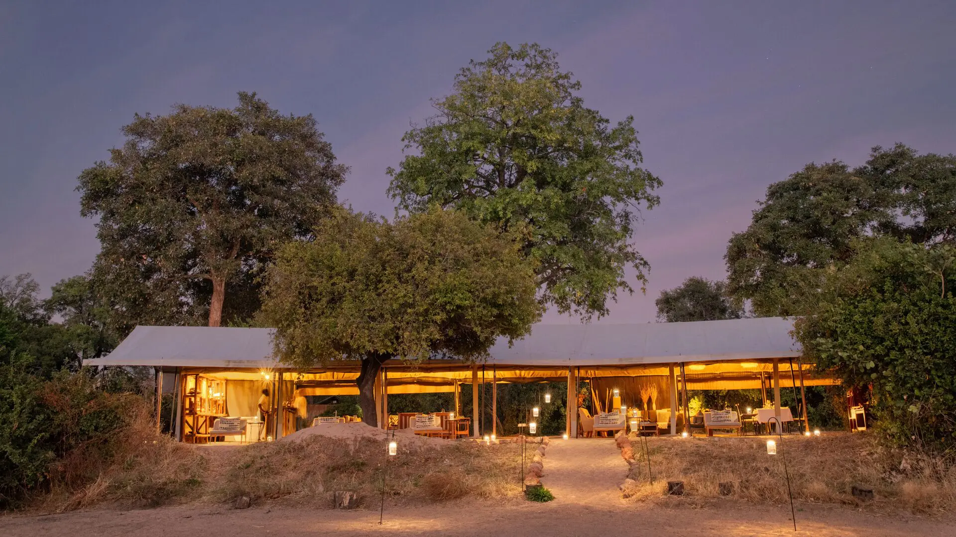 Kokoko Camp's softly lit lounge and dining area under canvas tents surrounded by trees at twilight.
