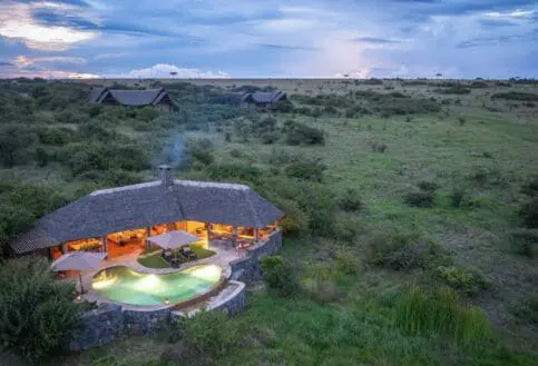 Aerial view of Naboisho Camp at dusk, featuring a lit main lodge with swimming pool, set within expansive green savannah under a dramatic evening sky.