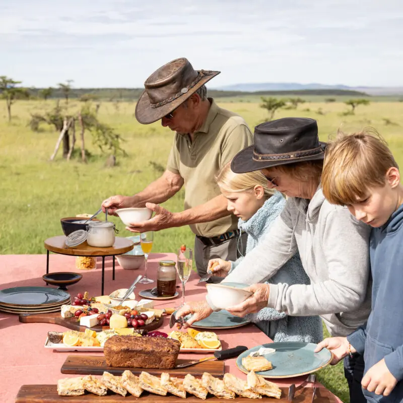 Guests and children serving themselves food at a bush breakfast table set in open grassland