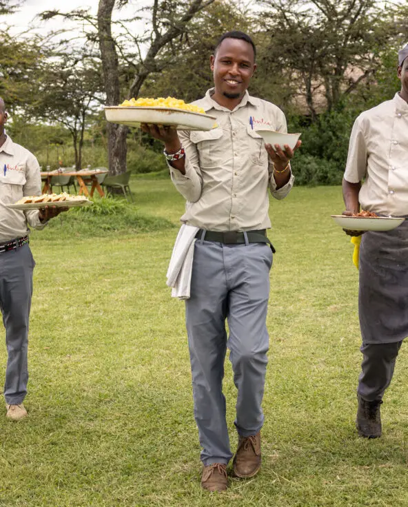 Three safari staff members walking across grass carrying plates of food toward guests