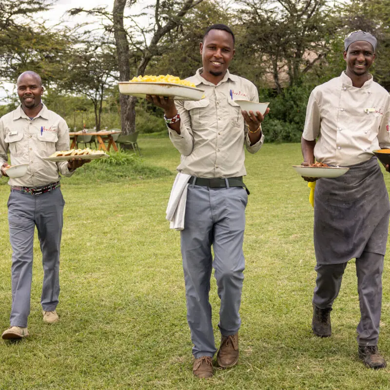 Three safari staff members walking across grass carrying plates of food toward guests
