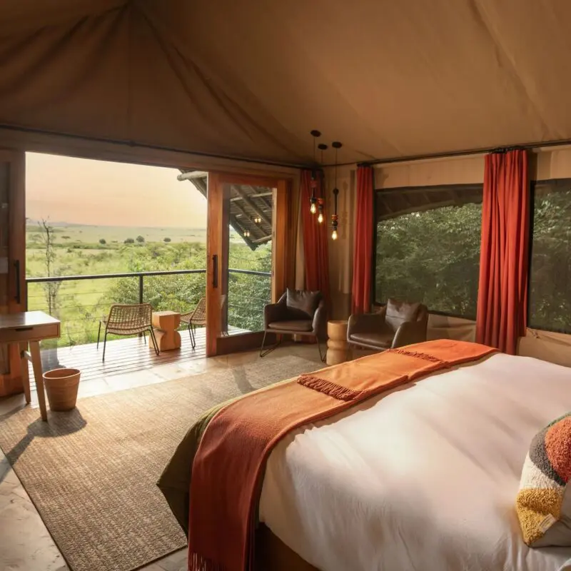 Interior of a luxury safari double room at Naboisho Camp with king bed, warm furnishings, large windows, and a private deck overlooking open savannah.