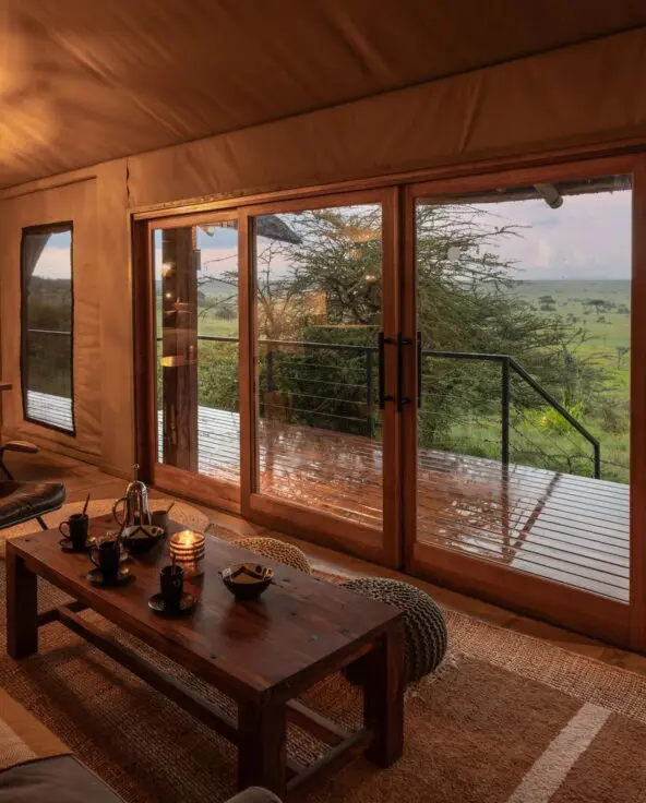 The family suite interior of Naboisho's luxury safari family suite with lounge and dining area, sliding glass doors, and a wooden deck overlooking expansive savannah views.
