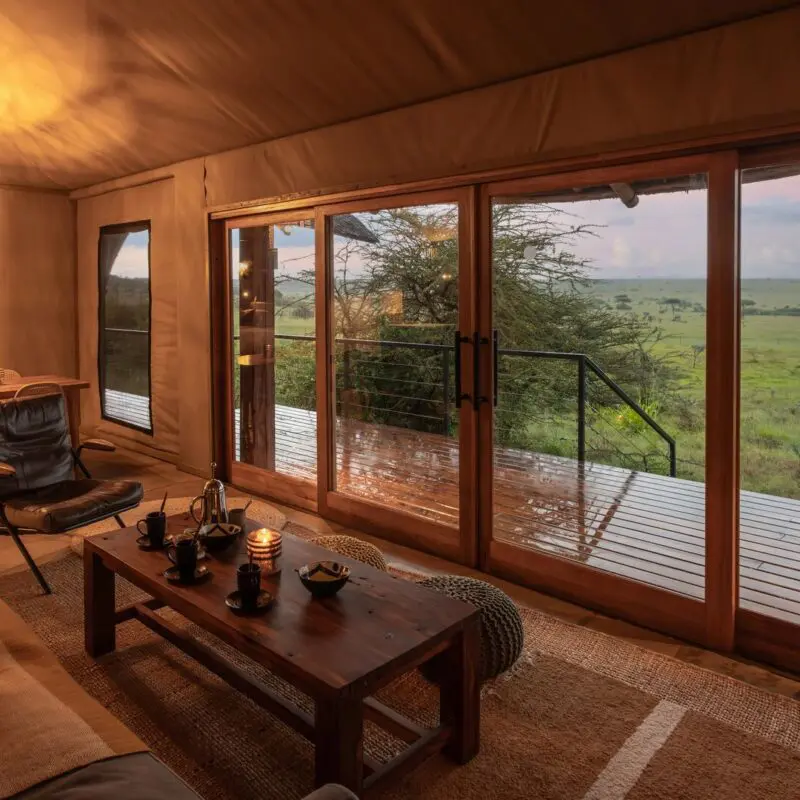 The family suite interior of Naboisho's luxury safari family suite with lounge and dining area, sliding glass doors, and a wooden deck overlooking expansive savannah views.