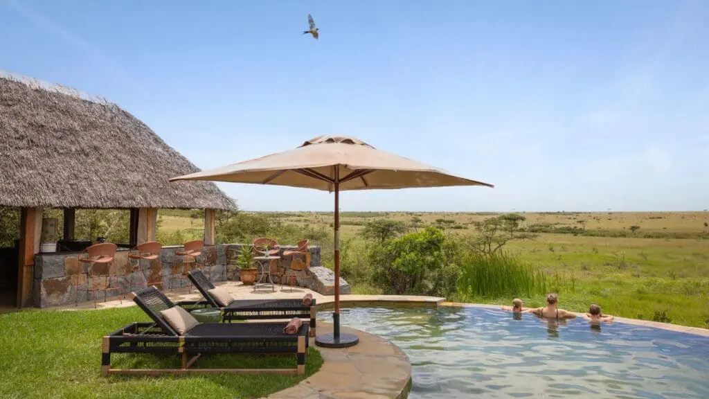 Guests enjoy the swimming pool at Naboisho Camp, with loungers, shaded seating, and wide-open savannah stretching into the distance under a clear sky.