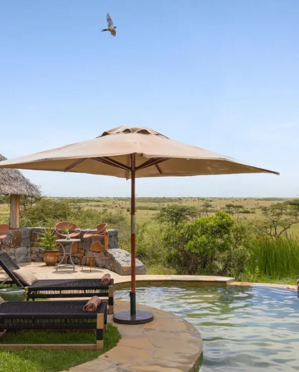 Guests enjoy the swimming pool at Naboisho Camp, with loungers, shaded seating, and wide-open savannah stretching into the distance under a clear sky.