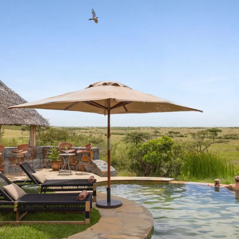 Guests enjoy the swimming pool at Naboisho Camp, with loungers, shaded seating, and wide-open savannah stretching into the distance under a clear sky.