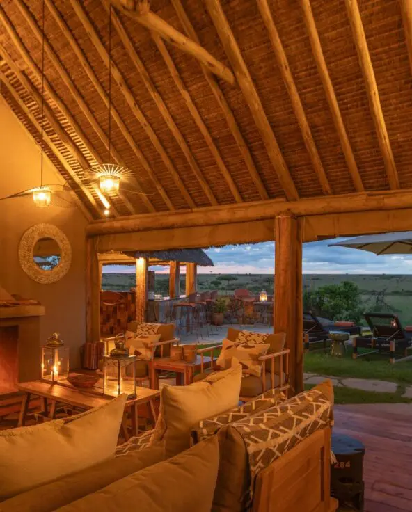 Naboisho Camp's lounge featuring a roaring fireplace, comfortable seating, wooden beams, and a deck overlooking the savannah and pool at dusk.