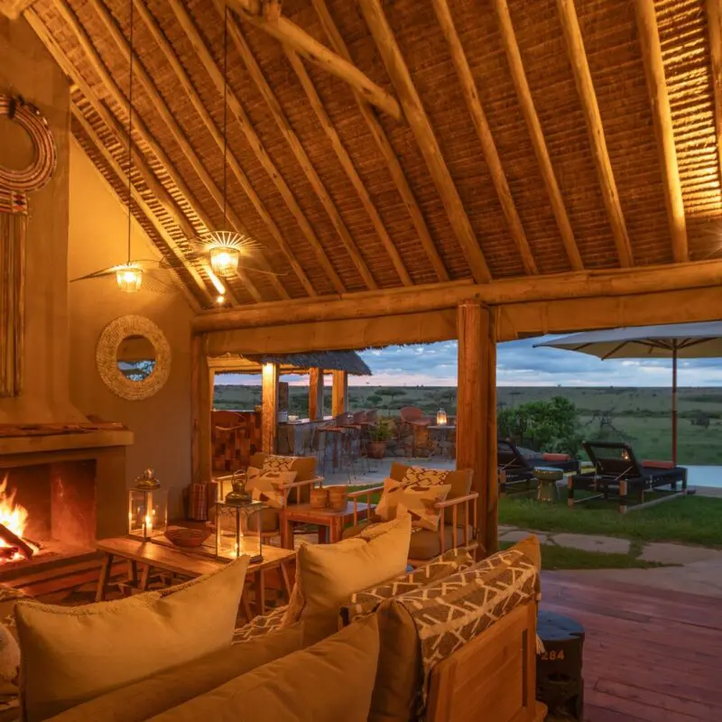 Naboisho Camp's lounge featuring a roaring fireplace, comfortable seating, wooden beams, and a deck overlooking the savannah and pool at dusk.