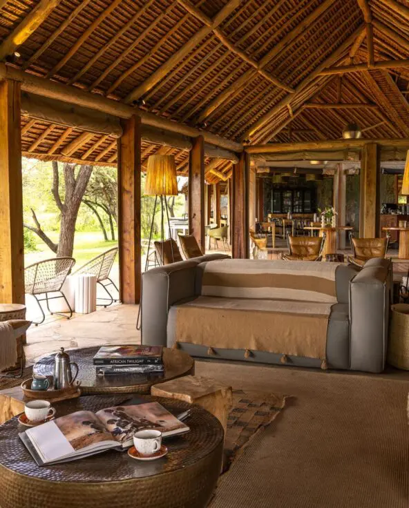 Naboisho Camp's open-sided lounge with comfortable seating, wooden beams, woven décor, and a bar area, opening onto greenery and shaded outdoor spaces.