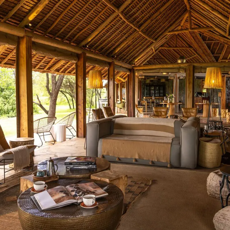 Naboisho Camp's open-sided lounge with comfortable seating, wooden beams, woven décor, and a bar area, opening onto greenery and shaded outdoor spaces.