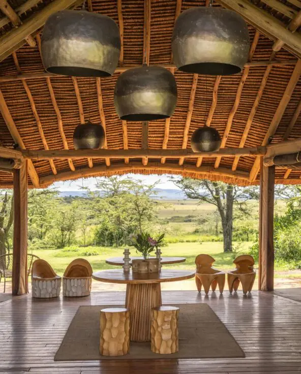 Naboisho Camp's main entrance with high thatched ceiling, wooden furnishings, sculptural seating, and open views across green savannah framed by acacia trees.
