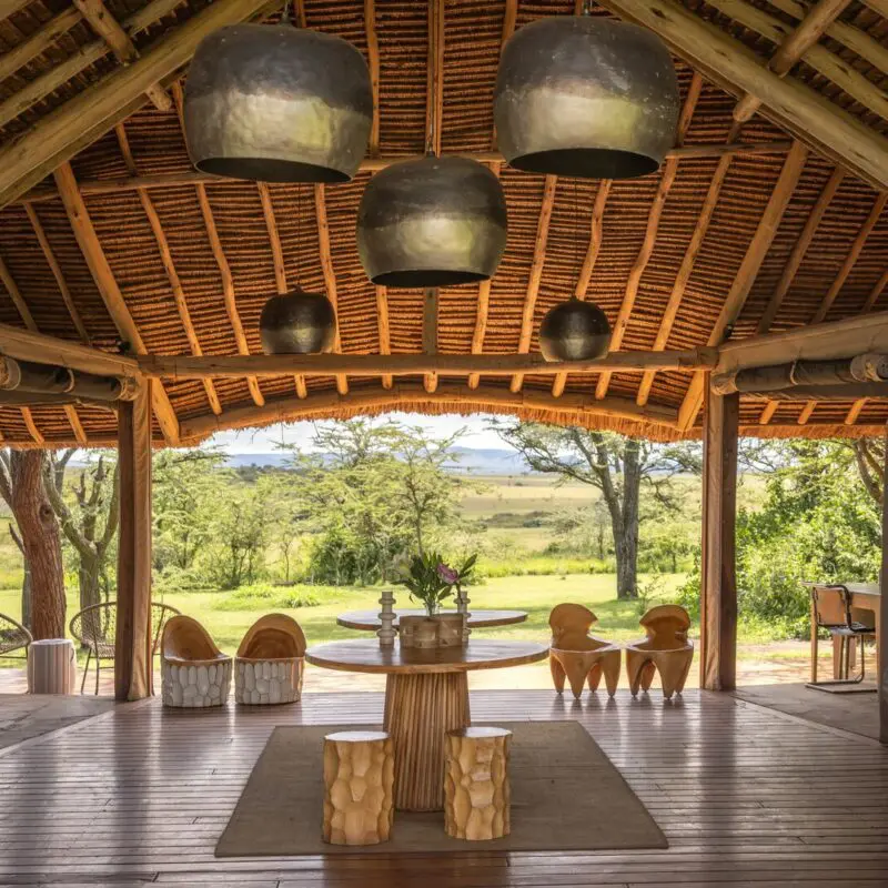 Naboisho Camp's main entrance with high thatched ceiling, wooden furnishings, sculptural seating, and open views across green savannah framed by acacia trees.