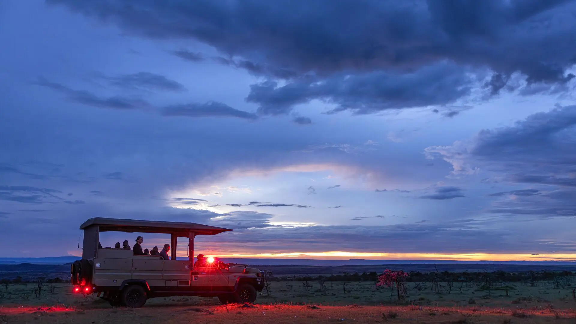 An open safari vehicle with guests overlooks the savannah on a night game drive at sunset under dramatic clouds