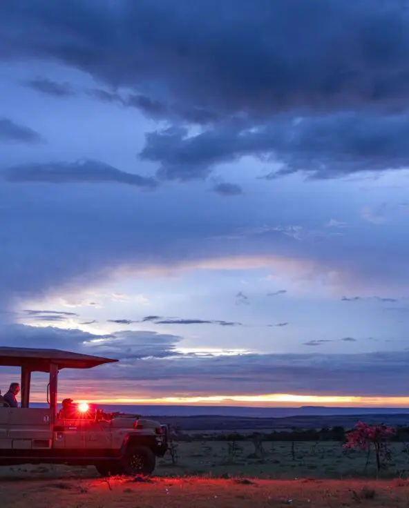 Guests ride in an open safari vehicle during a night game drive from Naboisho Camp, illuminated by red lights against a vast savannah and dramatic twilight sky.