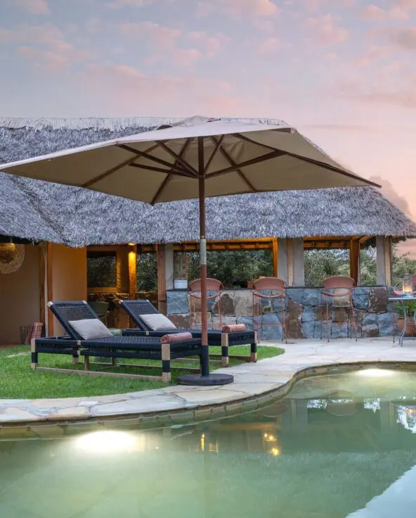 Naboisho Camp's stone-edged swimming pool, shaded loungers, and open lounge areas, set against expansive savannah views at sunset.