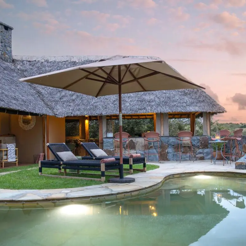 Naboisho Camp's stone-edged swimming pool, shaded loungers, and open lounge areas, set against expansive savannah views at sunset.