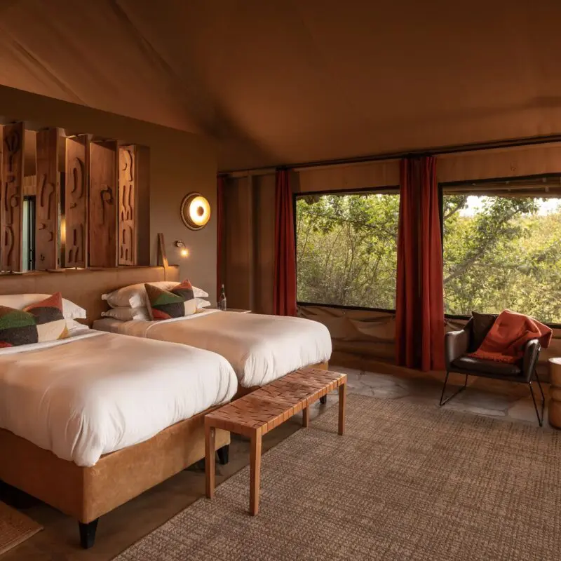Interior of a luxury twin at Naboisho camp room featuring two beds, warm lighting, woven textures, and large windows overlooking surrounding conservancy.