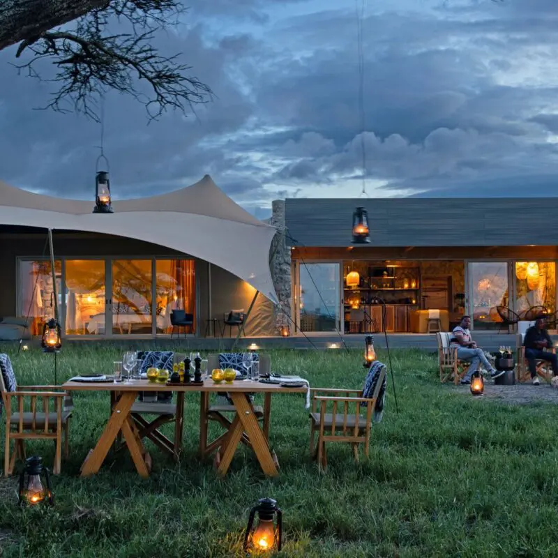Guests enjoy an outdoor evening meal and campfire at the Namiri Retreats with lanterns glowing, canvas tents illuminated, and dramatic clouds overhead.