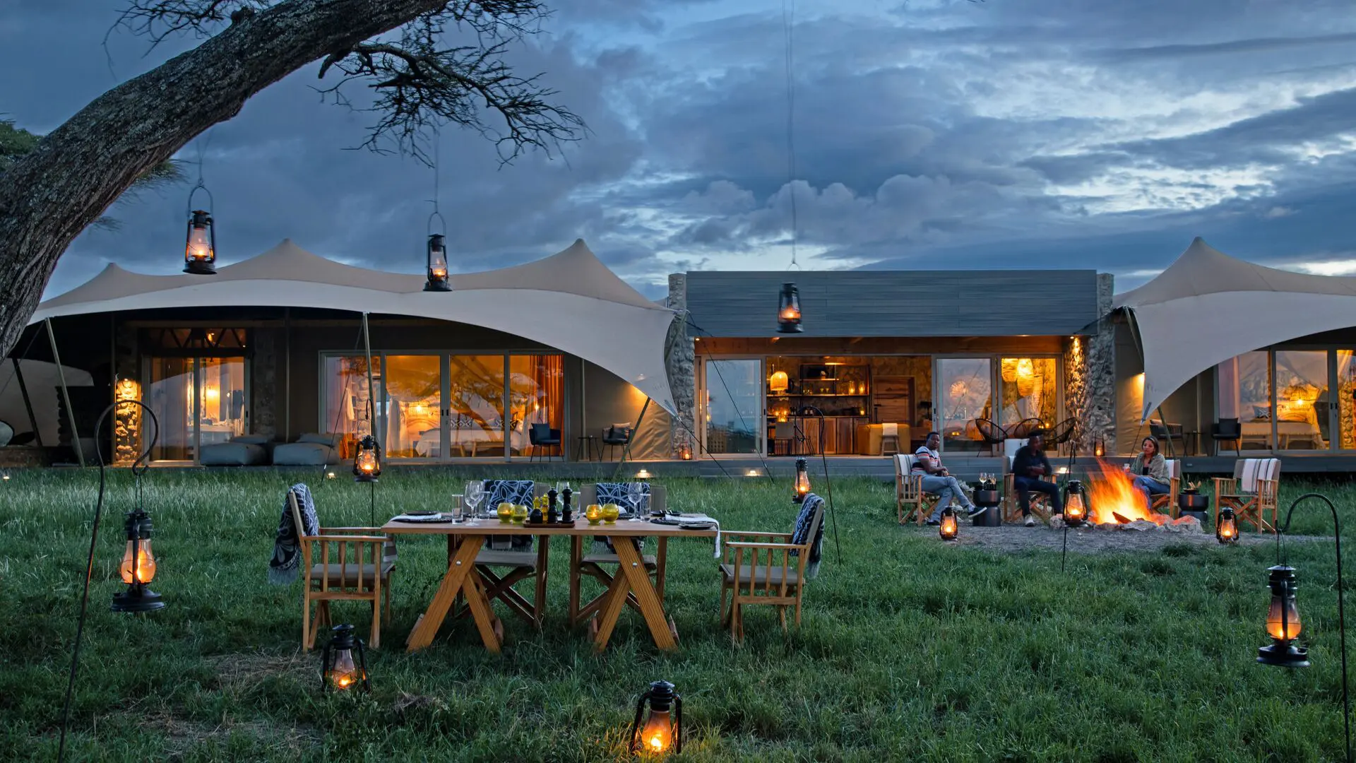 Guests enjoy an outdoor evening meal and campfire at the Namiri Retreats with lanterns glowing, canvas tents illuminated, and dramatic clouds overhead.