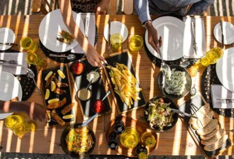 Overhead view of guests sharing a relaxed outdoor meal, with plated dishes, salads, bread, and drinks arranged on a wooden table in dappled sunlight.