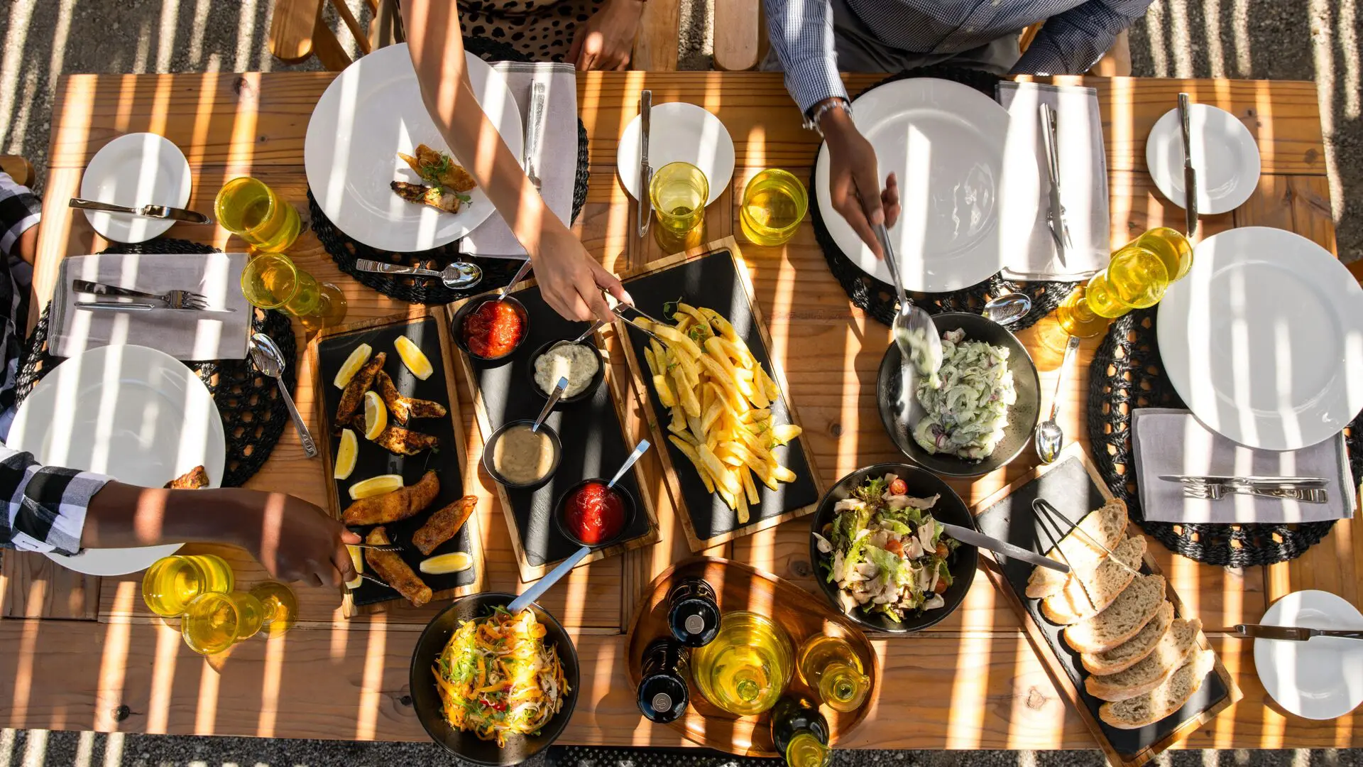 Overhead view of guests sharing a relaxed outdoor meal, with plated dishes, salads, bread, and drinks arranged on a wooden table in dappled sunlight.
