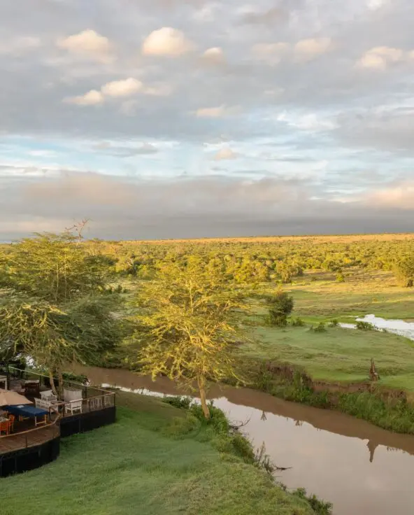 An aerial view of Ol Pejeta Safari camp set beside a winding river, with wooden decks nestled among trees and expansive green savannah stretching to the horizon under a soft, clouded sky.