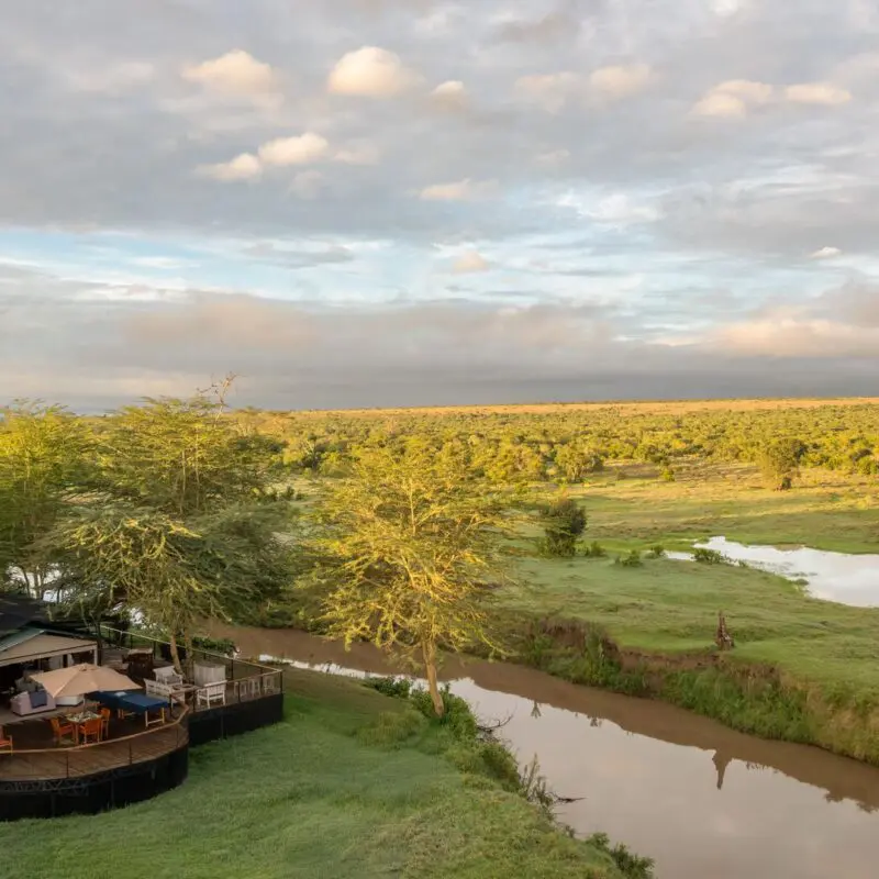 An aerial view of Ol Pejeta Safari camp set beside a winding river, with wooden decks nestled among trees and expansive green savannah stretching to the horizon under a soft, clouded sky.