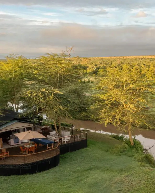 An aerial view of Ol Pejeta Safari camp set beside a winding river, with wooden decks nestled among trees and expansive green savannah stretching to the horizon under a soft, clouded sky.