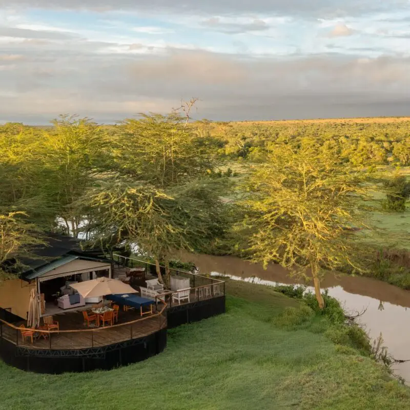 An aerial view of Ol Pejeta Safari camp set beside a winding river, with wooden decks nestled among trees and expansive green savannah stretching to the horizon under a soft, clouded sky.