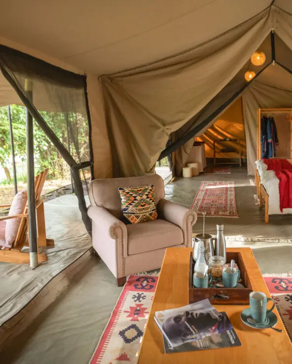 Interior of a large safari tent with seating area, beds, and open sides overlooking greenery