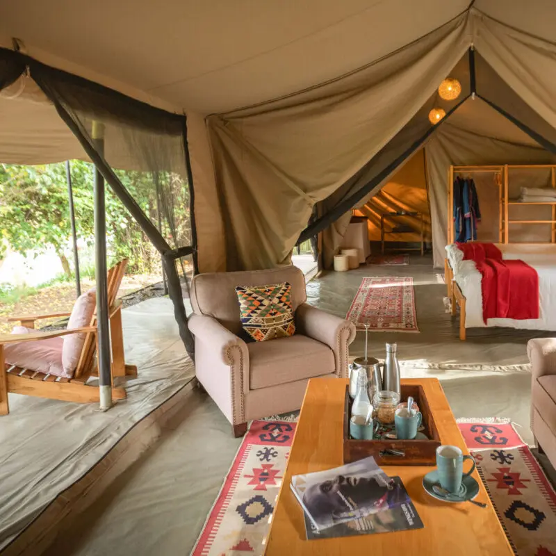 Interior of a large safari tent with seating area, beds, and open sides overlooking greenery
