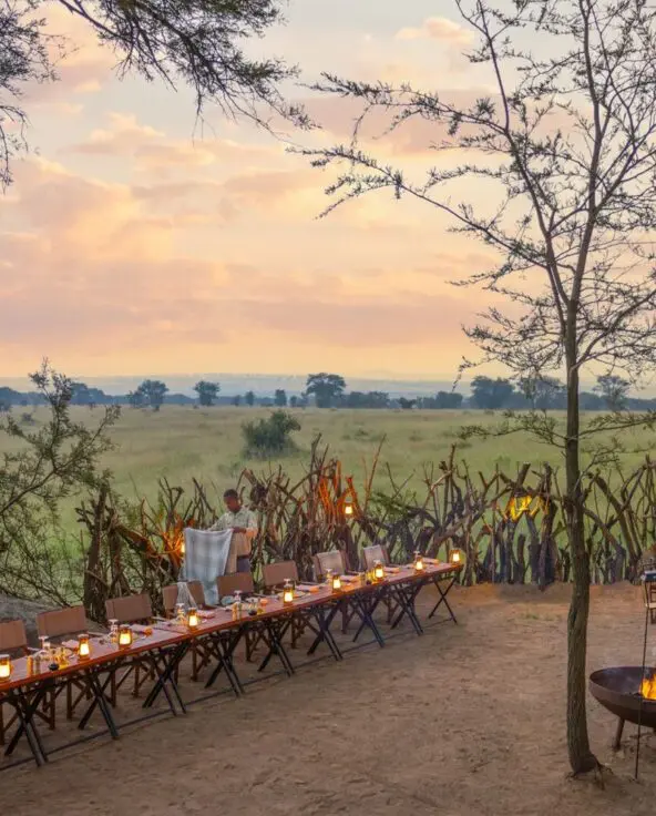 An outdoor safari dinner set at dusk, with long tables lit by lanterns, a small fire pit, rustic fencing, and expansive savannah views beneath a soft sunset sky.